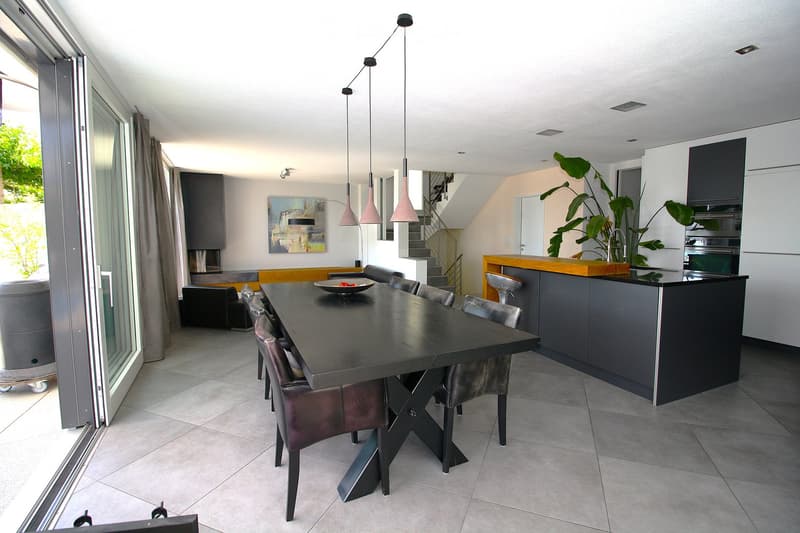 Modern dining area with a long black dining table surrounded by grey leather chairs, a sleek kitchen island with bar stools, pendant lights, and a large plant near the kitchen.