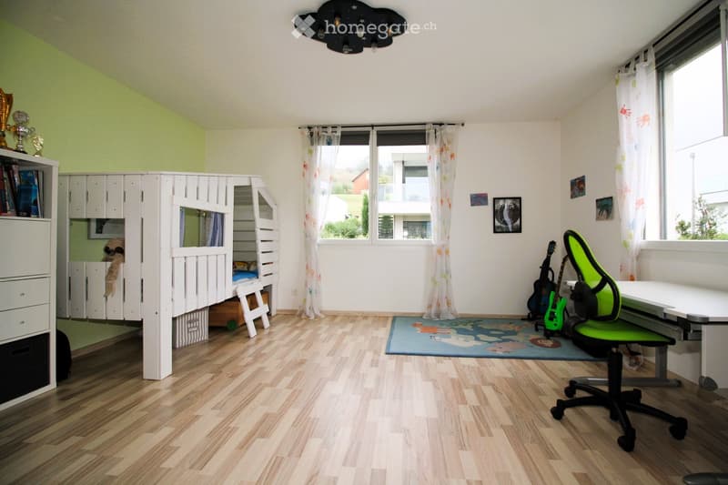 A well-lit room with wooden flooring, a bunk bed, a desk with a green chair, guitars, a colorful rug, and a bookshelf.