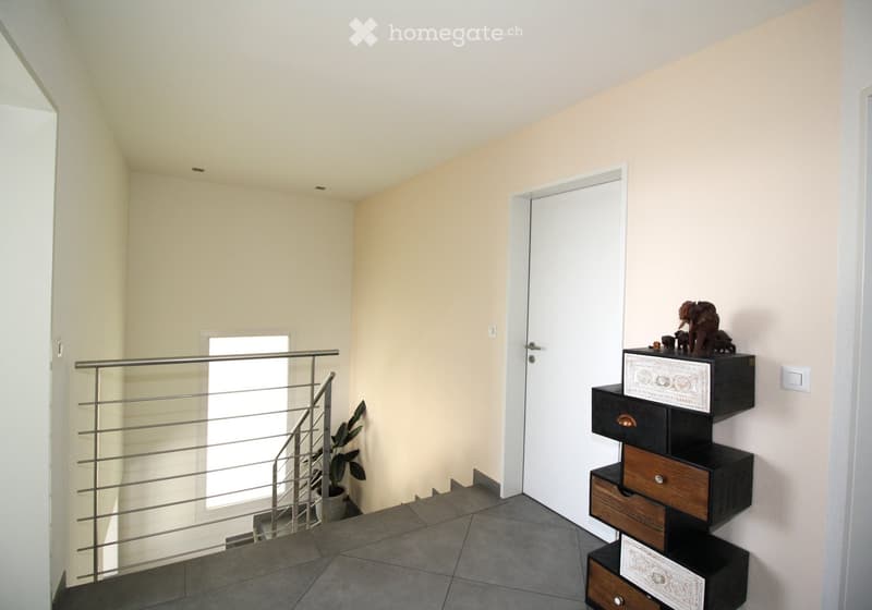 A modern hallway with a staircase, metal railing, a potted plant, and a decorative shelf with a statue on top.