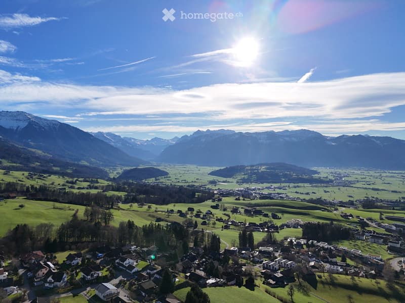 Aerial view of a picturesque village nestled in a lush green valley surrounded by snow-capped mountains. The village features numerous houses with varied roof styles, surrounded by fields and trees, under a clear blue sky with the sun shining brightly.