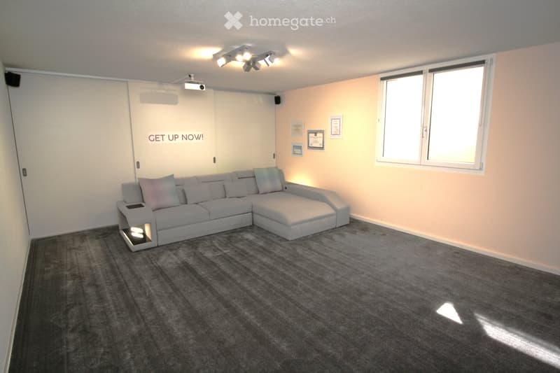 Modern living room with a grey sectional sofa, multiple pillows, a side table with built-in lighting, a projector mounted on the wall, and speakers. The room features large windows and a grey carpeted floor.