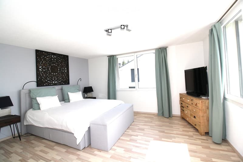 Modern bedroom with a large bed, nightstands, lamps, a TV on a wooden dresser, and large windows with curtains.