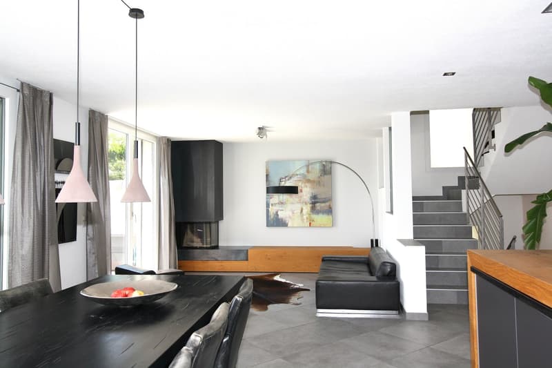 Modern living and dining area with a black leather sofa, dark wooden dining table, pendant lights, a large floor lamp, and a staircase leading to an upper level.