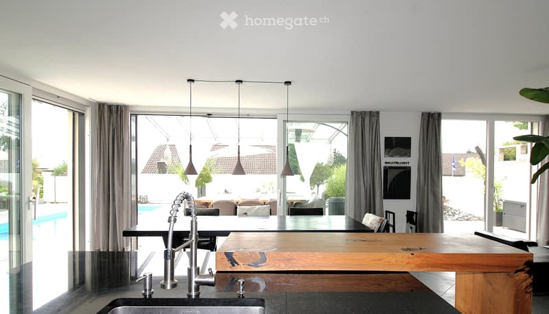 Modern kitchen with a wooden island, sink, and pendant lights, open to a dining area with a table and chairs, large windows overlooking a pool.