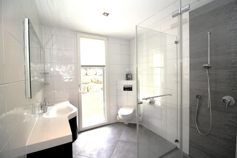 Modern bathroom with white tiles, a glass-enclosed shower, a sink with a mirror, and a toilet. The shower has a sleek, contemporary design with a handheld showerhead and a fixed showerhead. The bathroom features a large window that allows natural light to