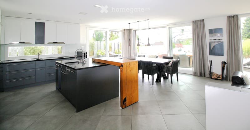 Modern kitchen with dark grey cabinets, a central wooden island, and a dining area with chairs and a table. Large windows with curtains provide natural light.