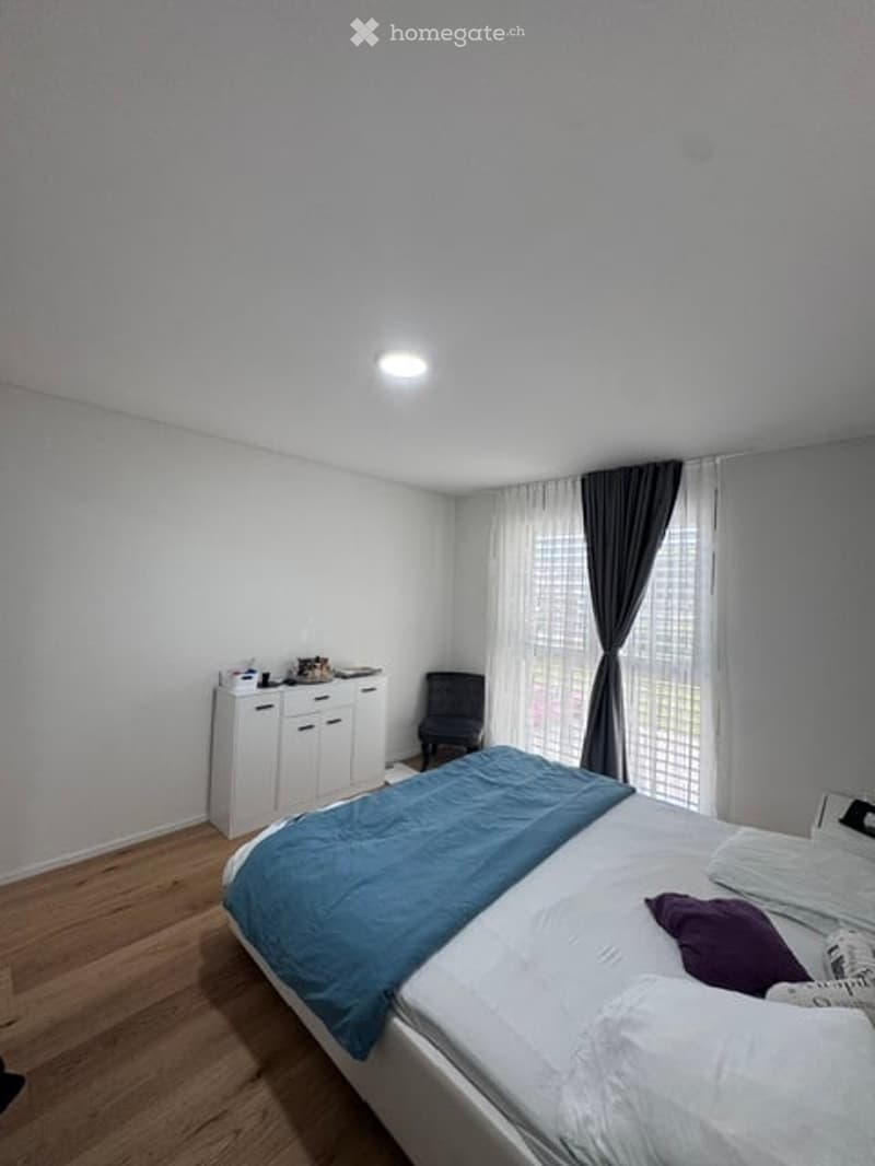 A modern bedroom with a large bed, a single chair, a small cabinet, and a window with curtains. The room features a wooden floor and white walls.