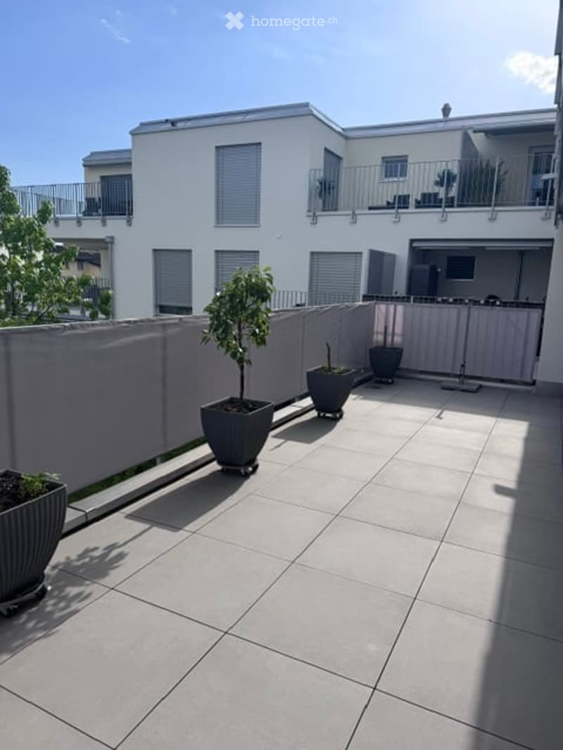 Modern balcony with tiled floor, grey planters, and a low fence. Adjacent building features balconies and large windows.