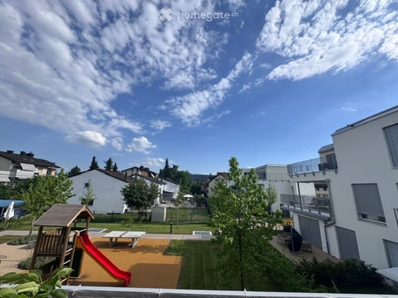 A well-maintained residential garden area with a playground, including a slide, climbing frame, and seating tables, surrounded by trees and neighboring houses.