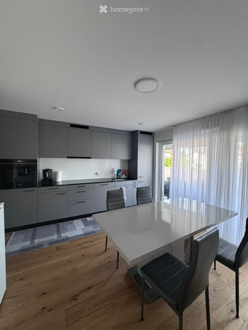 Modern kitchen with grey cabinets, a white dining table, and four chairs. A coffee maker is placed on the counter, and a large window with sheer curtains lets in natural light.