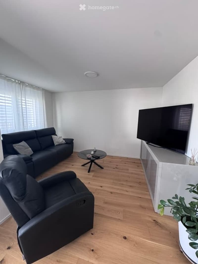 Modern living room with light wooden flooring, white walls, large windows with sheer curtains, black sofa, armchair, coffee table, flat-screen TV on a white stand, and a small plant on a side table.