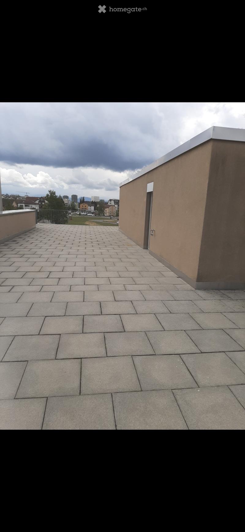 Empty rooftop terrace with tiled floor, low walls, and a view of the city and surrounding landscape.