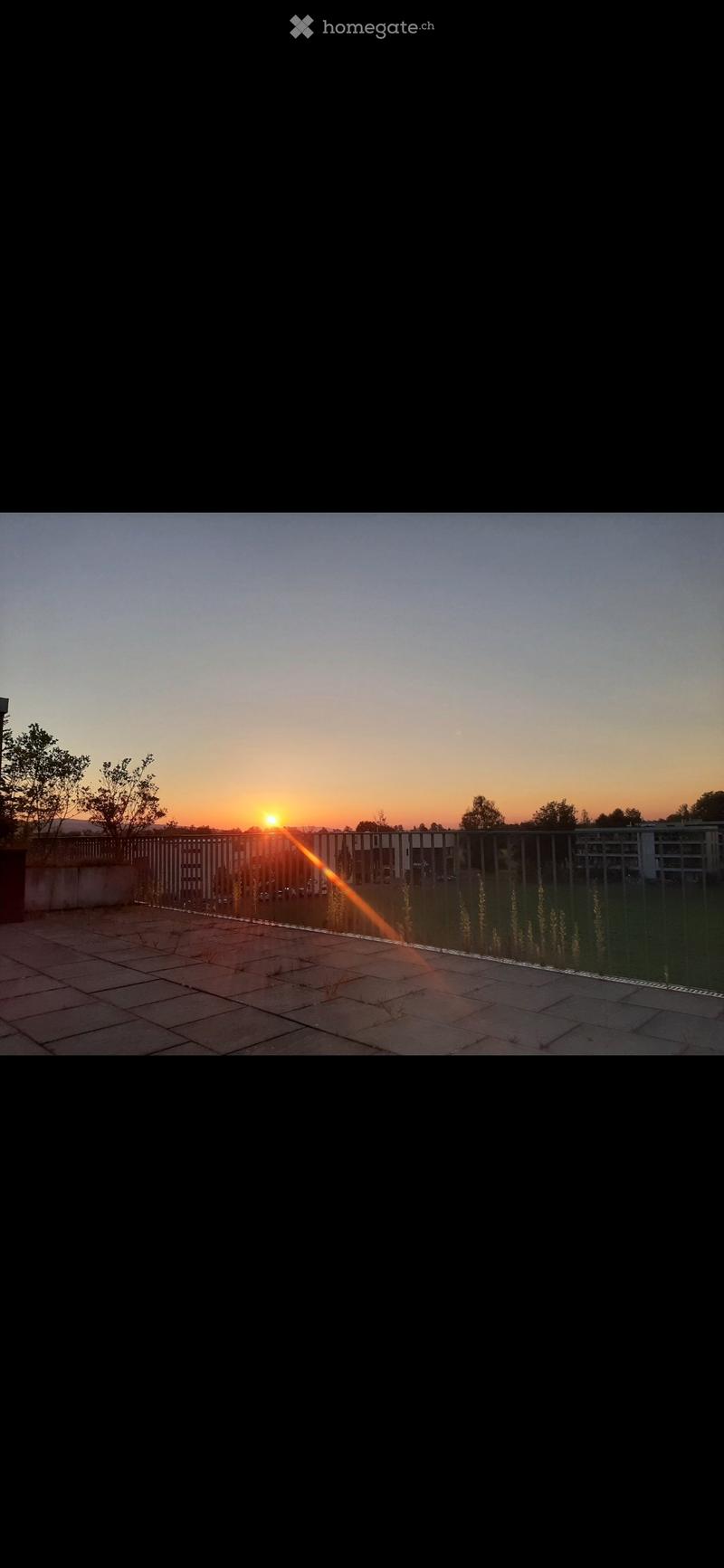 Balcony with tiled floor, metal railing, and a view of the sunset over a cityscape with buildings and trees.