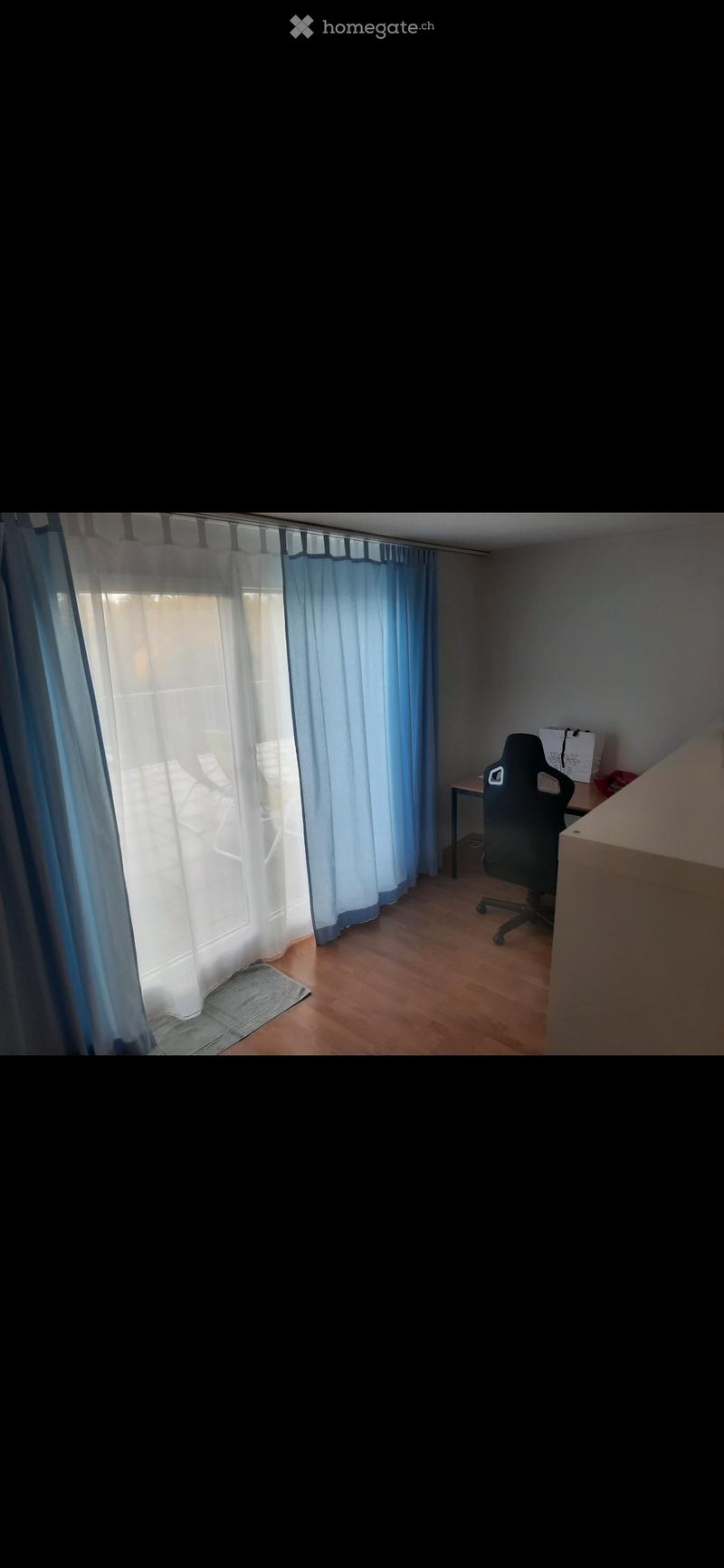 A small, furnished room with a wooden floor, a window with blue curtains, a chair, and a small table.