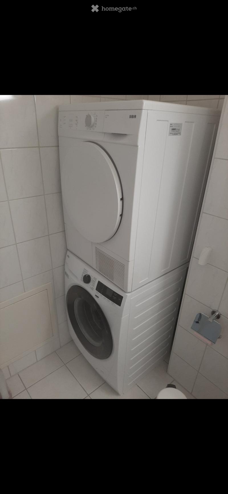 A compact bathroom with a stacked washing machine and dryer unit, white tiled walls and floor, and a small shelf with a folded towel.