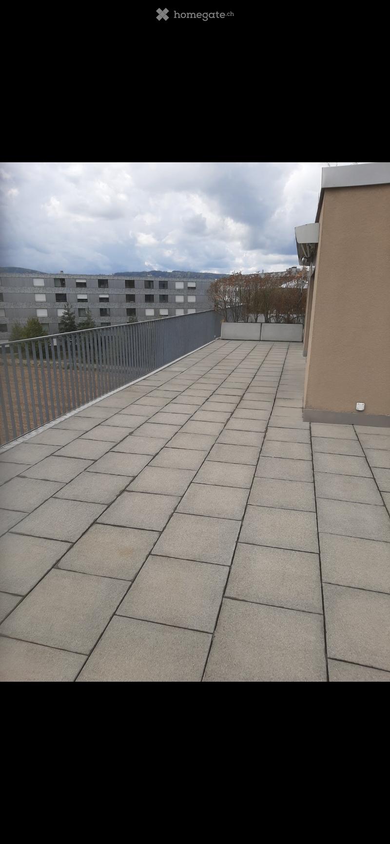 Empty balcony with tiled floor, metal railing, and a view of a building and trees in the background.