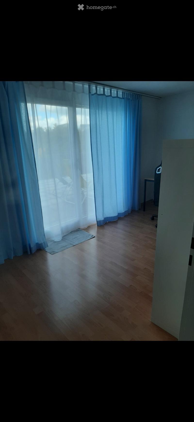 Empty room with wooden flooring, large windows covered by light blue curtains, and a small rug near the window. No furniture or equipment is present.