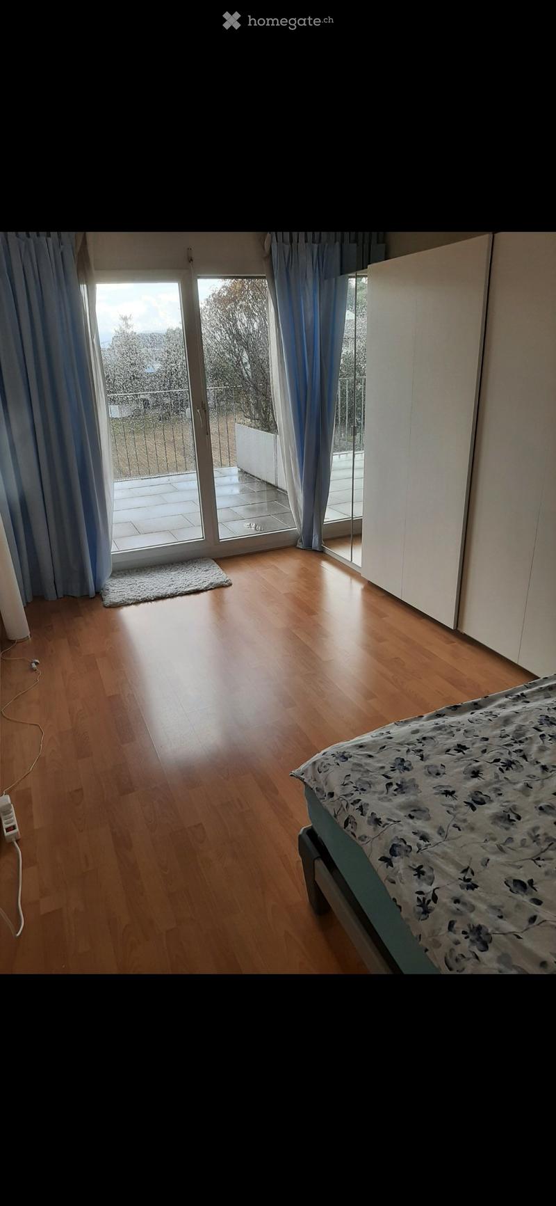 Bright bedroom with wooden flooring, large sliding glass doors leading to a balcony, a bed with floral bedding, and a white wardrobe.