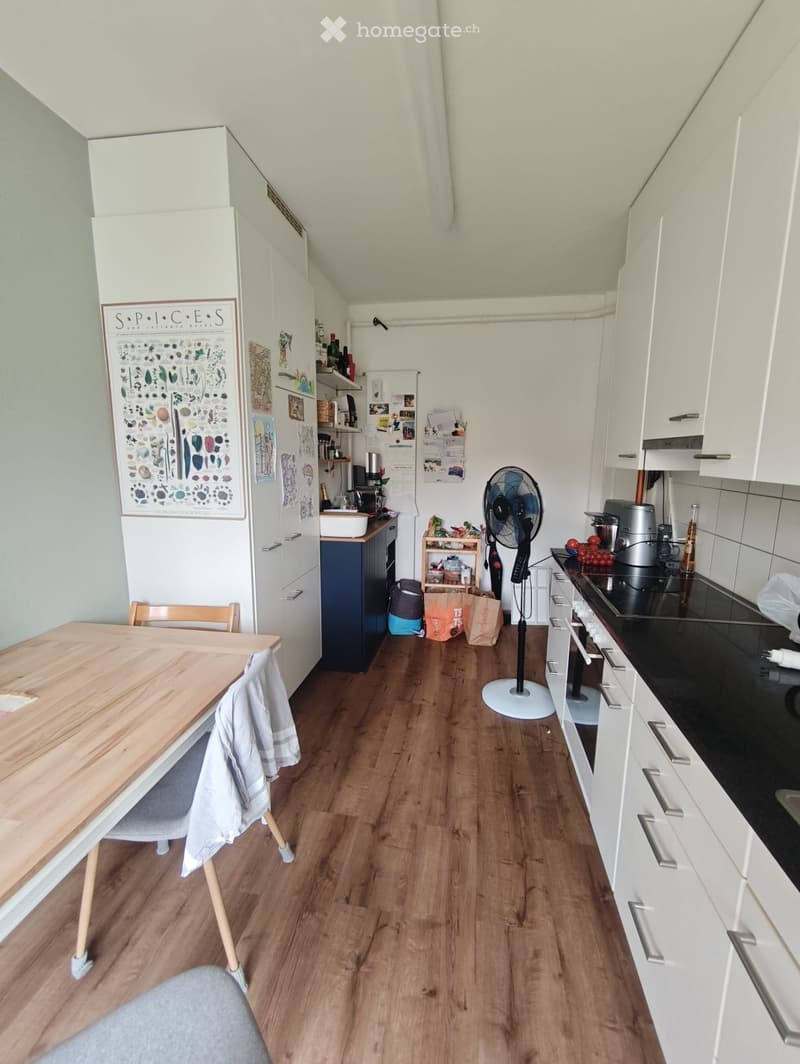 Modern kitchen with white cabinets, wooden flooring, a small dining table with a chair, a standing fan, and various appliances including a refrigerator and oven.