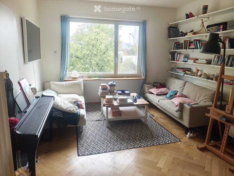 Living room with wooden flooring, large window with blue curtains, two sofas, coffee table with books, bookshelf, and a piano.