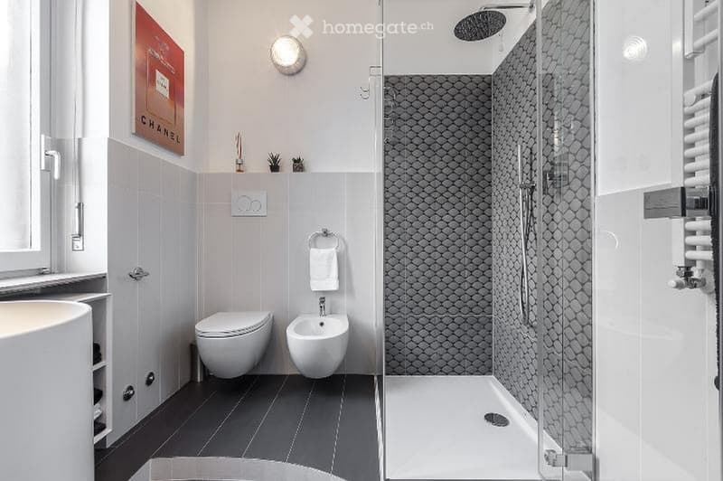 Modern bathroom with a glass-enclosed shower, dark tiled floor, white fixtures including a sink, toilet, and bidet, and a towel hanging on a ring. A radiator is visible on the right side, and a picture hangs on the wall.