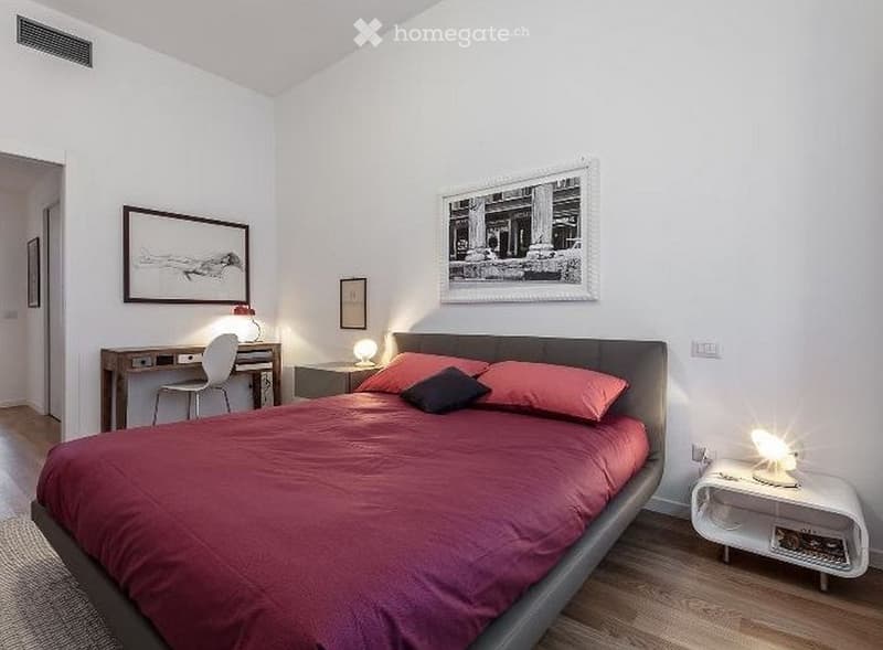 Modern bedroom with a large bed, red bedding, nightstand, desk, and chair. The room features wooden flooring and several framed artworks on the walls.