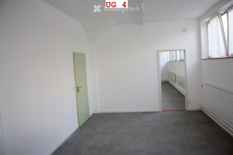 Empty room with white walls, grey carpeted floor, a single green door, and a window with white frame and curtains. The room is connected to another space through an open doorway.