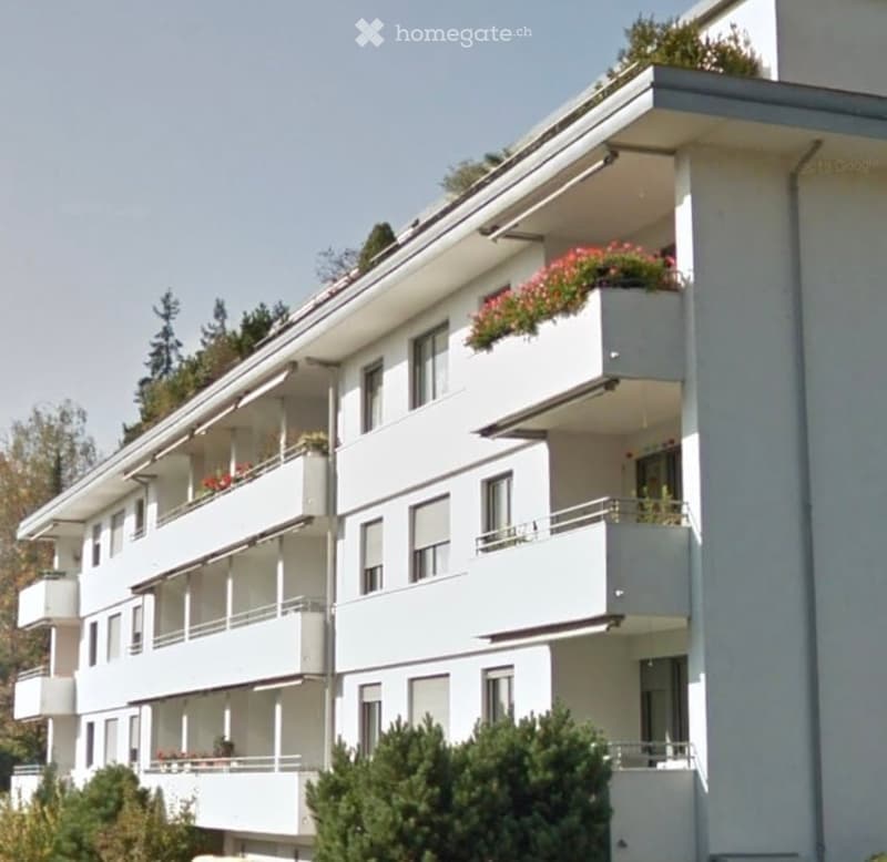 Modern multi-story apartment building with white balconies and flower boxes, surrounded by greenery.