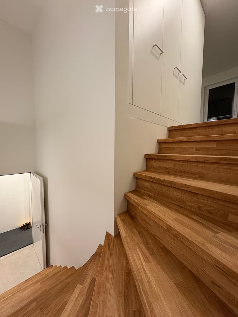 Wooden staircase with light-colored steps and railings, white walls, and a glass door leading to another room.