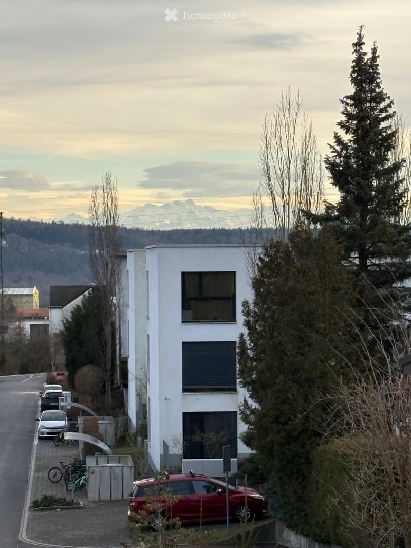 Modern white building with large windows, surrounded by trees and parked cars, with a scenic mountain view in the background.