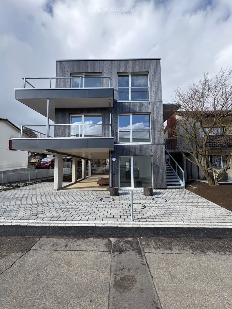 Modern two-story house with grey wooden facade, large windows, balconies, and a covered parking area.