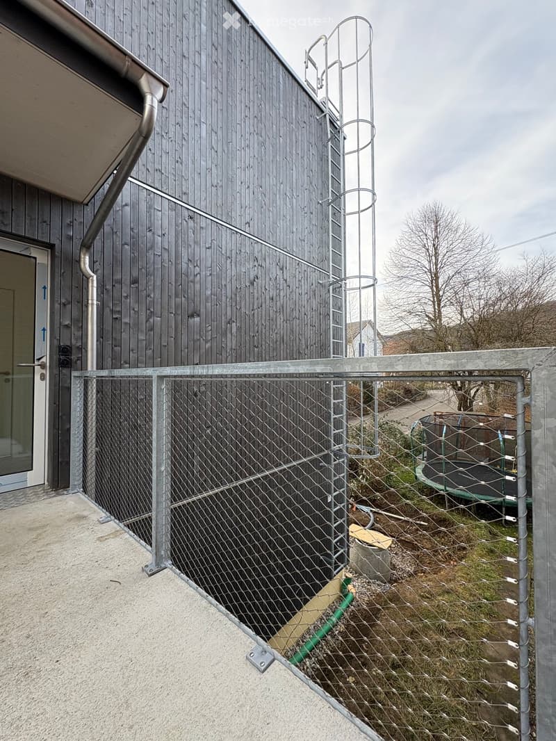 Outdoor area enclosed by a metal fence, with a modern building featuring wooden siding and a metal spiral staircase in the background. A trampoline is visible beyond the fence.