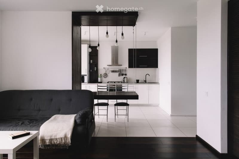 Modern open-plan living and dining area with a black sofa, coffee table, dining table with chairs, and a sleek kitchen area with cabinets, appliances, and a sink.