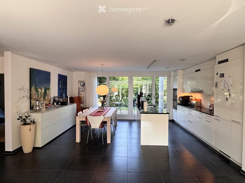 Open-plan kitchen and dining area with white cabinets, dark tiled floor, pendant light, dining table with chairs, and a view to the garden.