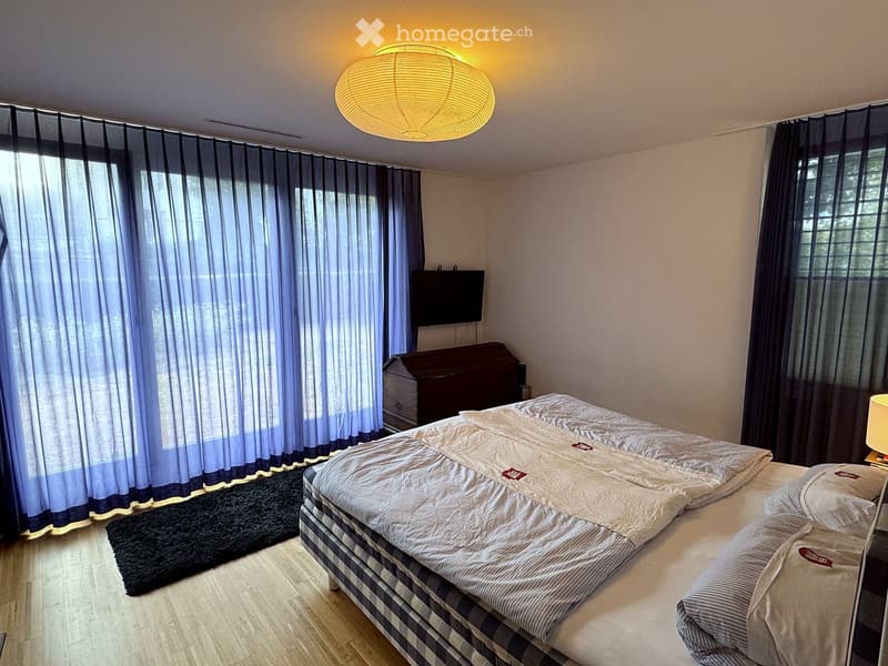 Modern bedroom with a large double bed, nightstand, lamp, and television mounted on the wall. The room features wooden flooring, large windows with dark curtains, and a sleek, contemporary design.