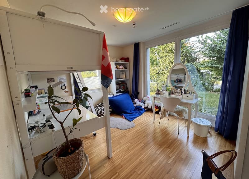 A cozy, multi-functional room with a lofted bed, desk, and sitting area. Features wooden flooring, large windows with curtains, and a variety of furniture including a desk, chair, mirror, and potted plant.