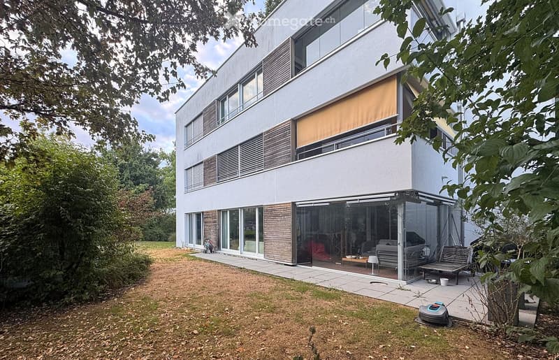 Modern two-story house with white facade, large windows, and a covered outdoor patio area. The house features a sleek design with wooden accents and a well-maintained garden.
