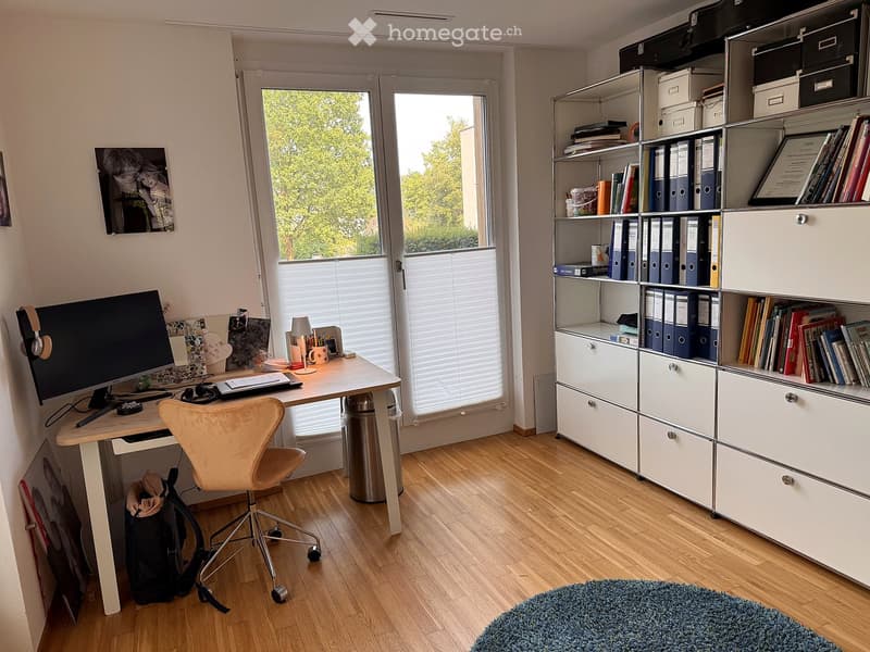 Well-lit study room with a wooden floor, large window with blinds, a modern desk setup, ergonomic chair, and a tall shelving unit filled with books and files.