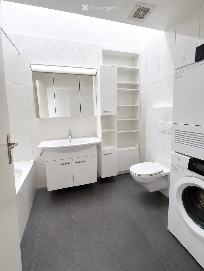 Modern bathroom with white tiles, a sink, a toilet, a washing machine, a bathtub, and a mirrored cabinet.