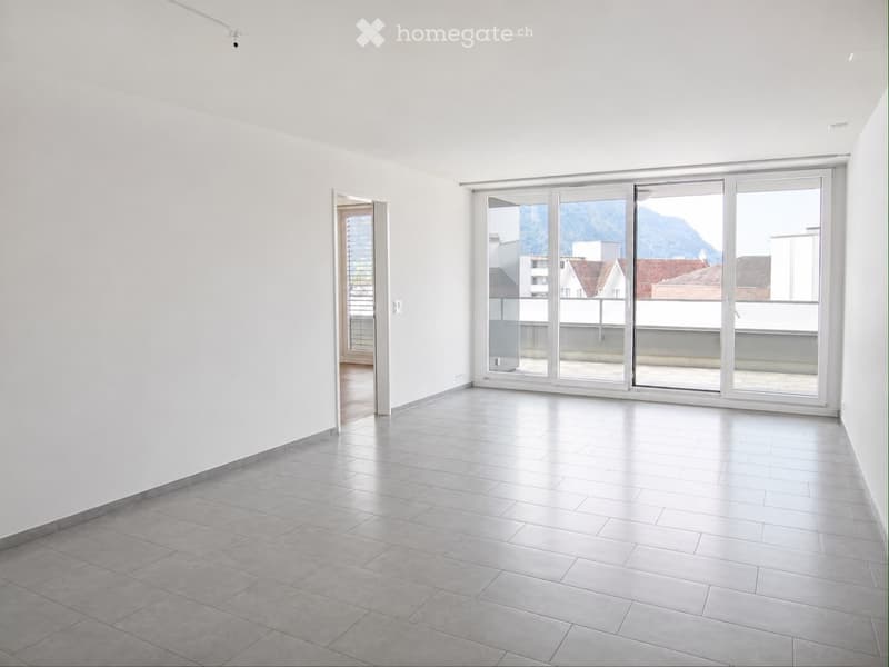 Empty room with white walls, grey tiled floor, large sliding glass doors leading to a balcony, and a view of mountains and buildings.