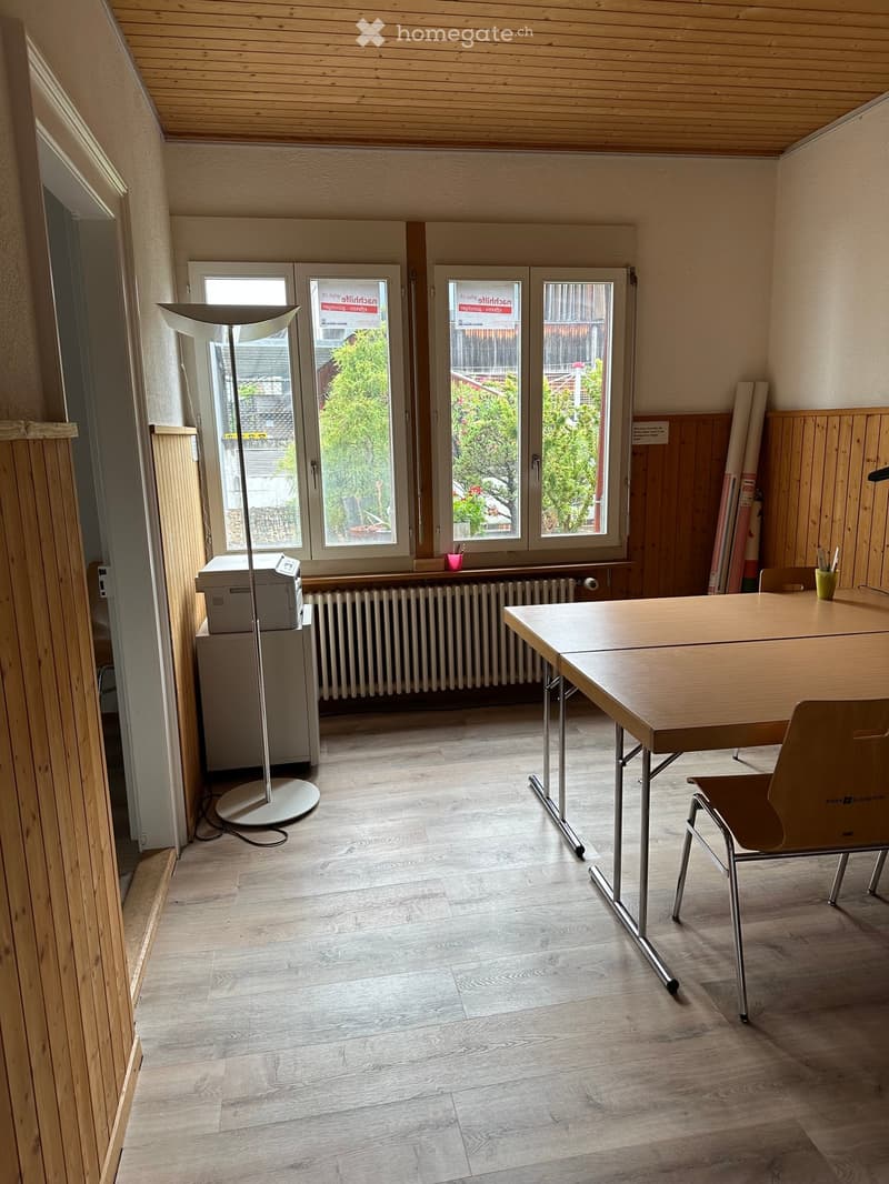 Bright room with wooden flooring, large windows, a table with chairs, a floor lamp, and a printer on a stand.