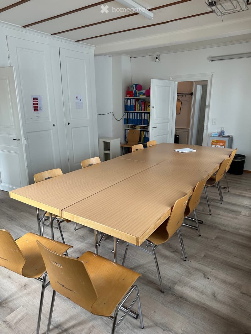 A furnished room with a long wooden table surrounded by multiple chairs, a bookcase with books and binders, and a small shelf unit.