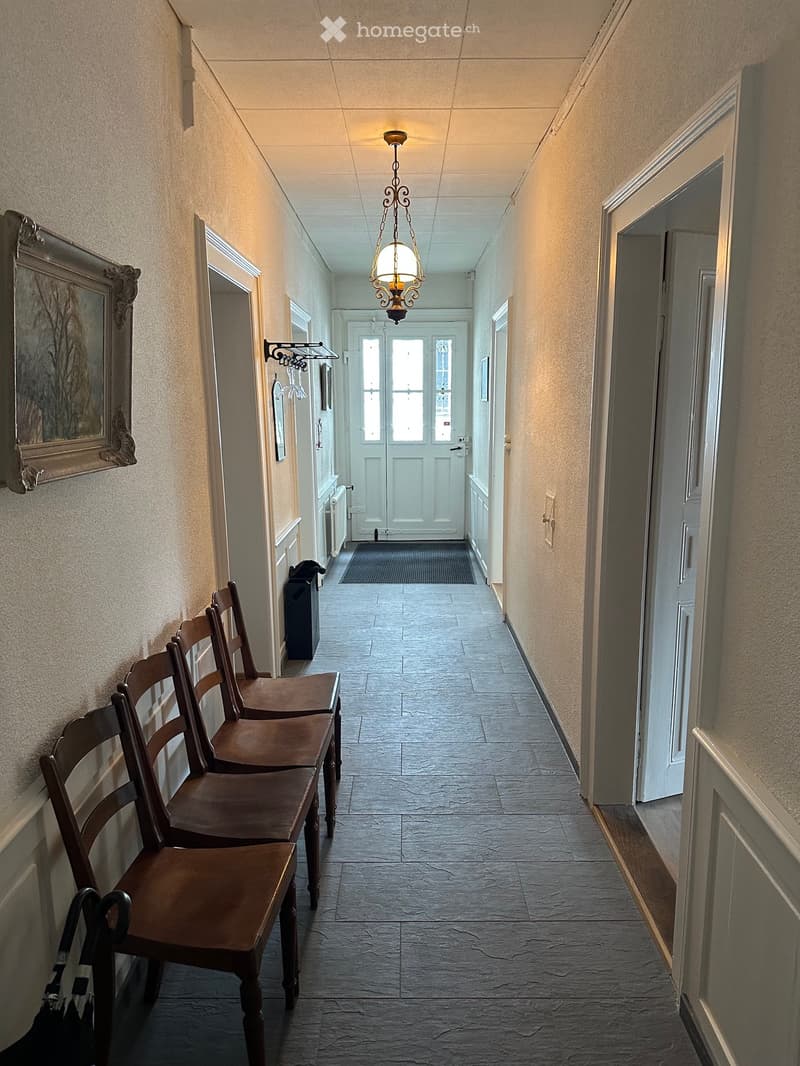 Hallway with beige walls, tiled floor, wooden chairs, a chandelier, and a framed picture on the wall.
