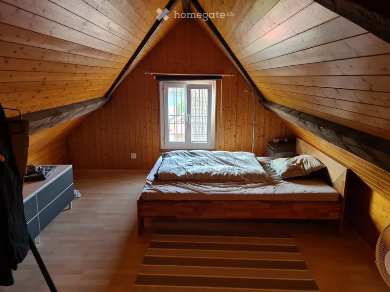 Cozy attic bedroom with wooden walls and slanted ceilings, featuring a single bed, nightstand, and a fan near the window.