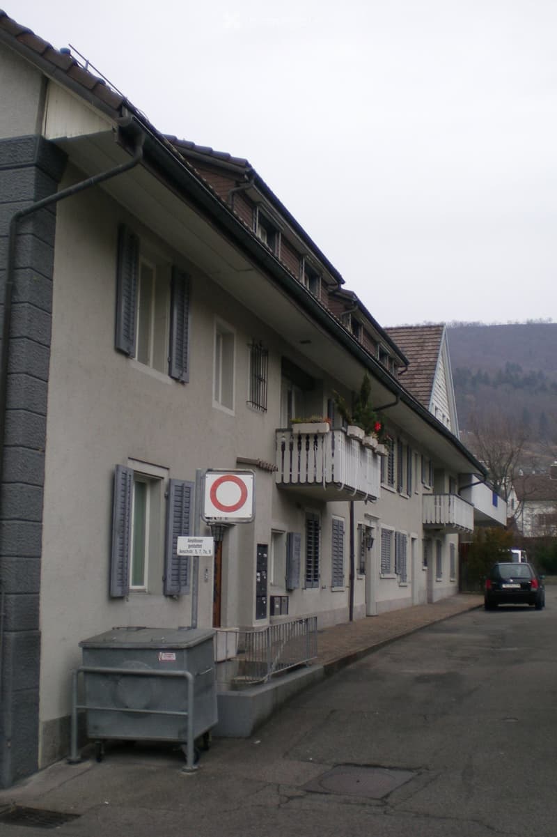 Multi-story residential building with white facade, multiple windows, balconies, and a trash container on the sidewalk.
