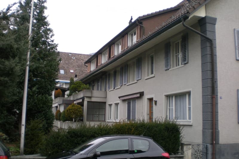 A multi-story residential building with a gray facade, multiple windows with shutters, and a small balcony on the upper floor. The building is surrounded by greenery and has a parked car in the foreground.