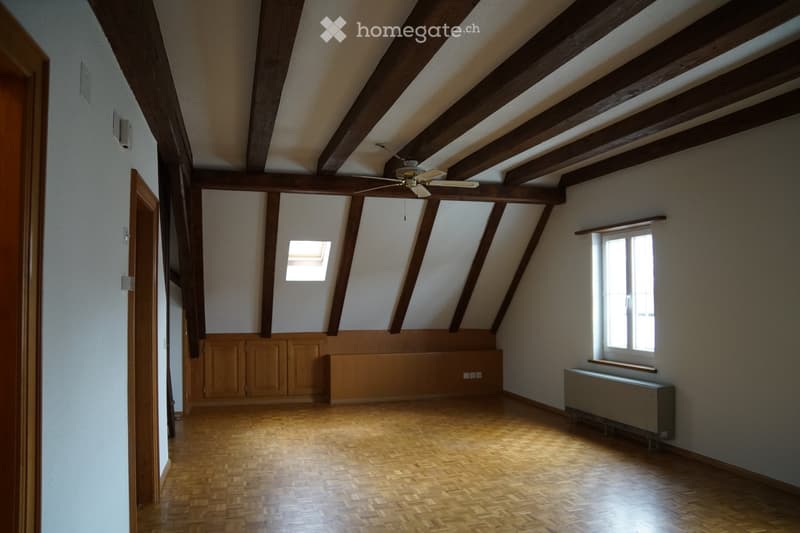 Empty attic room with wooden beams, exposed wooden ceiling, wooden floor, and two windows allowing natural light. Features include a radiator, a small skylight, and a ceiling fan.