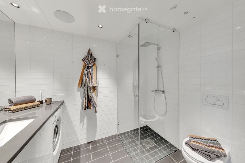 Modern bathroom with white tiled walls, a glass-enclosed shower, a sink with a mirror, a toilet, and a washing machine. Towels and a bathrobe are neatly arranged on the counter and toilet seat.