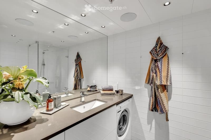 Modern bathroom with white tiles, double sink, large mirror, towel rack, and a washing machine. Features a decorative plant and toiletries on the counter.