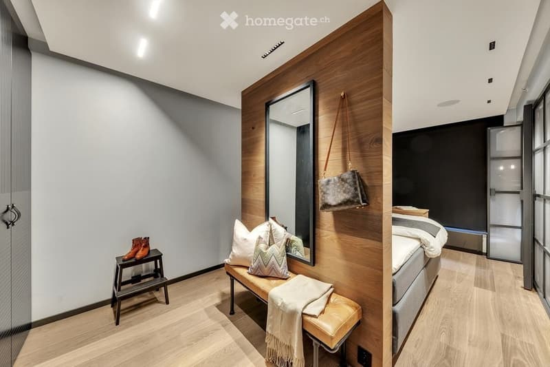 Modern bedroom with wooden flooring, a large mirror, a bench with pillows and a blanket, a small stool with shoes, and a bed with pillows and a blanket.